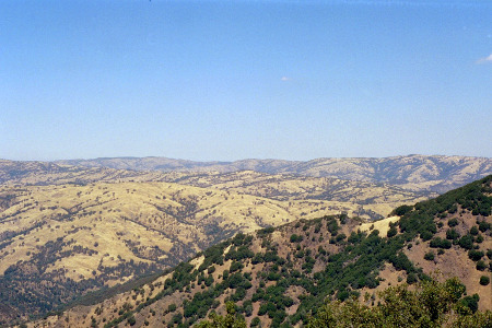 [This image is taken near the top and is mostly dirt with scattered sections of trees. The hills are smooth rather than rocky and jagged and softened by the haze of the distance except for the ones closest to the camera. The sky only has a couple of wisps of clouds.]